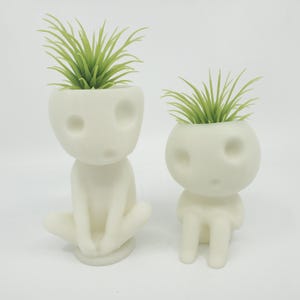 Puede incluir: Dos macetas de cer&aacute;mica blanca con forma de peque&ntilde;as figuras humanas con plantas verdes que crecen de sus cabezas.