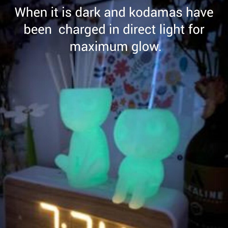 Puede incluir: Dos figuras de kodama verdes brillantes con pajitas, sentadas sobre una superficie de madera con un reloj digital que muestra las 7:31. El texto "When it is dark and kodamas have been charged in direct light for maximum glow." es visible sobre las figuras.