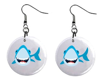 May include: A pair of silver dangle earrings with a white circular charm featuring a cartoon blue shark with a toothy grin.