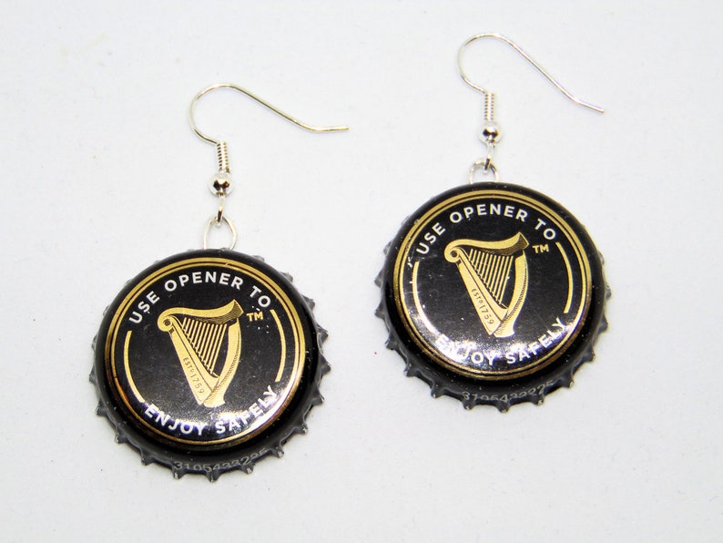 May include: A pair of earrings made from black and gold Guinness bottle caps. The caps have the text "Use opener to enjoy safely" and a harp logo.