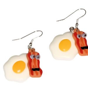 May include: A pair of dangle earrings featuring a white fried egg with a yellow yolk and a red and orange bacon strip with googly eyes and a mustache.