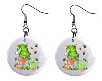 May include: A pair of earrings featuring a gnome holding carrots and a watering can. The gnome is wearing a green hat and has a white beard. The earrings are surrounded by bees.