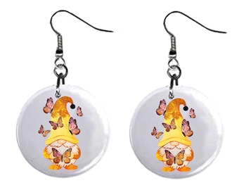 May include: A pair of dangle earrings featuring a gnome with a yellow hat and a butterfly in its hands. The gnome is surrounded by butterflies with purple and orange wings. The earrings are on a white background.