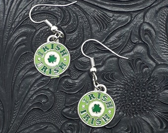 May include: A pair of silver earrings with green and white enamel charms. The charms are round and feature the word "Irish" written in a circle around a shamrock.