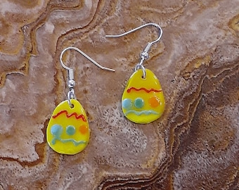 May include: A pair of yellow Easter egg earrings with red, orange, and blue accents. The earrings are made of glass and have silver hooks.