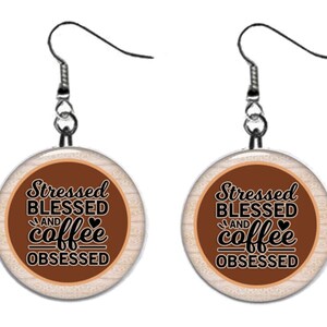 May include: A pair of silver earrings with brown and white round pendants. The pendants have a wood-grain texture and feature the text "Stressed Blessed and coffee obsessed" in black.