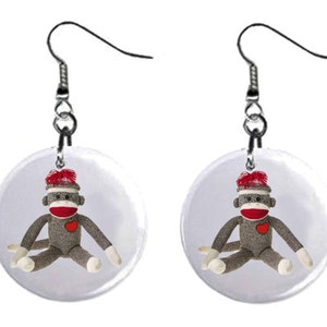 May include: A pair of earrings featuring a round, clear button with a sock monkey design. The monkey is wearing a red hat and has a red heart on its chest.