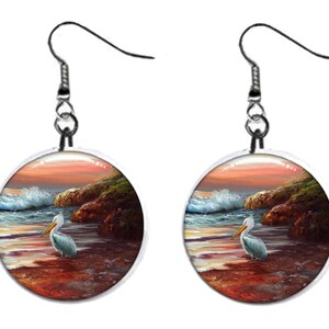 May include: A pair of round earrings featuring a coastal scene with a pelican standing on the shore. The image depicts a sunset with waves crashing against the rocks. The earrings have silver-colored hooks.