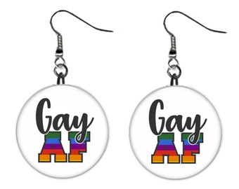 May include: A pair of silver dangle earrings with round white button charms. The charms have the text "Gay AF" in black with a rainbow colored "AF".