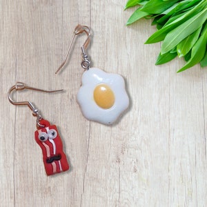 May include: A pair of earrings featuring a fried egg and a cartoon bacon slice. The egg earring is white with a yellow yolk, while the bacon earring is red with a white stripe and a black mustache.