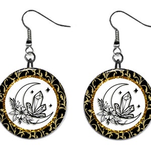 May include: A pair of silver dangle earrings with black and gold round charms. The charms feature a crescent moon with a crystal and a floral design on a white background.