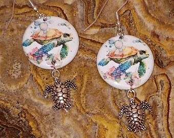 May include: A pair of silver dangle earrings with a white round charm featuring a watercolor painting of a sea turtle swimming in a blue ocean with bubbles. The charm is attached to a silver turtle charm.