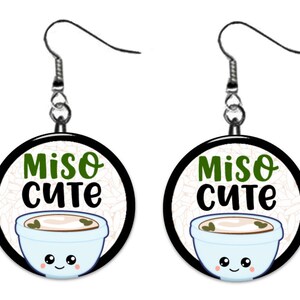 May include: A pair of round earrings with a black border. Each earring features a light blue bowl of miso soup with a smiling face, and the words "Miso Cute" in green and black. The background has a rice pattern.