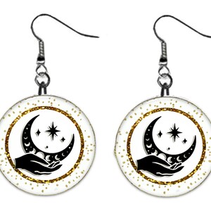 May include: A pair of silver dangle earrings with a white round charm featuring a black silhouette of a hand holding a crescent moon and stars on a gold glitter background.