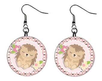 May include: A pair of silver dangle earrings with pink and brown circular charms featuring a cartoon hedgehog with closed eyes and pink flowers.