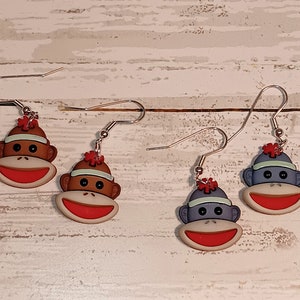 May include: Three pairs of monkey-themed earrings. Each pair features a different colored monkey with a red flower on its head. The monkeys are wearing a blue, green, or red hat.
