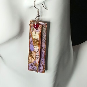 May include: A pair of dangle earrings with a layered design. The earrings are made of copper and feature a purple, gold, and white patterned paper. The earrings have a red bead at the top.
