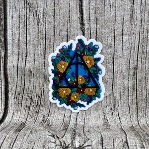 Peut inclure: Un autocollant représentant une illustration en noir et blanc du symbole des Reliques de la Mort, entourée d'une couronne de fleurs colorées. Les fleurs sont jaunes, rouges et vertes.
