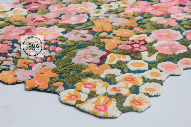 Pu&ograve; includere: Un tappeto floreale con sfondo turchese e un motivo di fiori colorati nelle tonalit&agrave; del rosa, arancione e bianco. Il tappeto ha un bordo ritagliato e un logo di "The Rug Company".