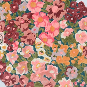 Pu&ograve; includere: Un tappeto floreale con sfondo verde acqua e un motivo di fiori colorati nelle tonalit&agrave; del rosa, arancione e bordeaux. Il tappeto ha un bordo smerlato e un logo nell'angolo.