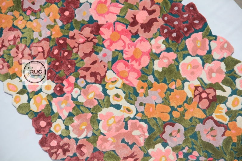 Pu&ograve; includere: Un tappeto floreale con sfondo verde acqua e un motivo di fiori colorati nelle tonalit&agrave; del rosa, arancione e bordeaux. Il tappeto ha un bordo smerlato e un logo nell'angolo.