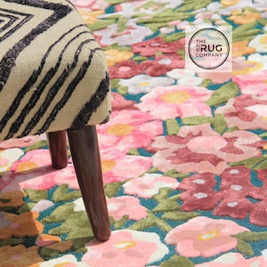 Pu&ograve; includere: Un tappeto floreale con sfondo turchese e fiori rosa, rossi e gialli. Una sedia beige e nera a fantasia &egrave; in primo piano. Il logo The Rug Company &egrave; visibile.