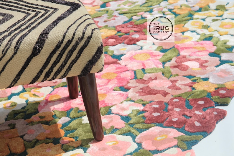 Pu&ograve; includere: Un tappeto floreale con sfondo turchese e fiori rosa, rossi e gialli. Una sedia beige e nera a fantasia &egrave; in primo piano. Il logo The Rug Company &egrave; visibile.