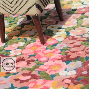 Pu&ograve; includere: Un tappeto floreale con sfondo verde acqua e un motivo di fiori rosa, rossi e gialli. Il tappeto &egrave; parzialmente coperto dalle gambe di una sedia in legno con seduta fantasia. Il logo The Rug Company &egrave; visibile.