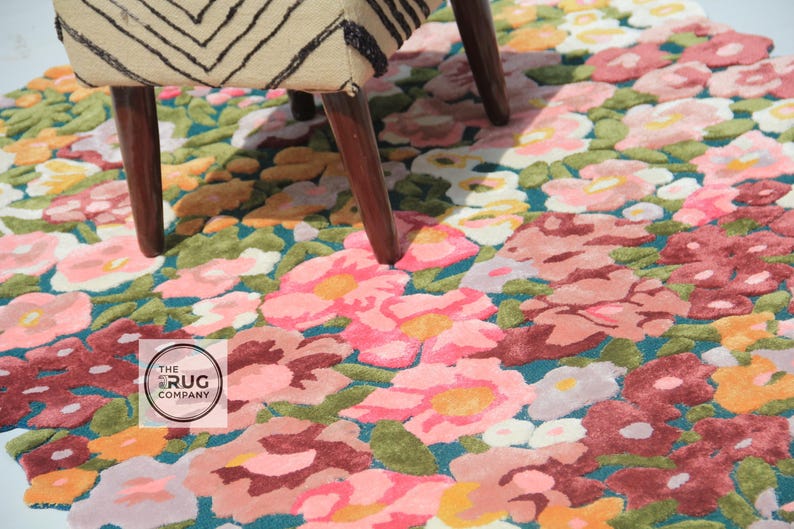 Pu&ograve; includere: Un tappeto floreale con sfondo verde acqua e un motivo di fiori rosa, rossi e gialli. Il tappeto &egrave; parzialmente coperto dalle gambe di una sedia in legno con seduta fantasia. Il logo The Rug Company &egrave; visibile.