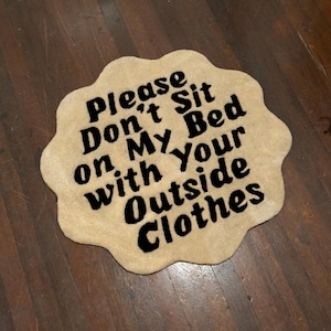 Pode incluir: Um tapete bege em forma de nuvem com uma borda ondulada. O tapete apresenta o texto "Please Don't Sit on My Bed with Your Outside Clothes" em letras pretas em negrito. O tapete está colocado em um chão de madeira.