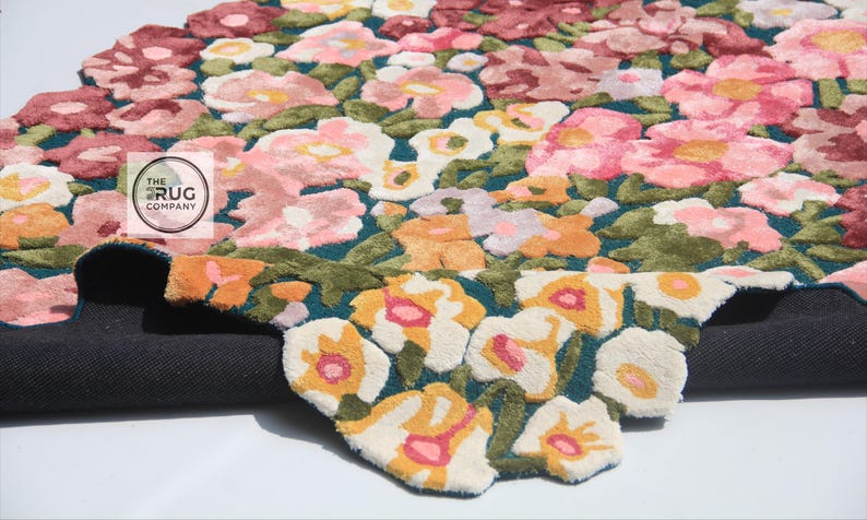Pu&ograve; includere: Un tappeto floreale con sfondo verde acqua scuro e un motivo di fiori rosa, pesca e bianchi. Il tappeto ha una trama a pelo tagliato e un retro grigio scuro. Il logo The Rug Company &egrave; visibile.