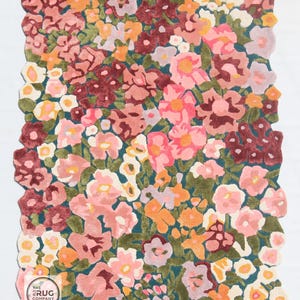 Pu&ograve; includere: Un tappeto floreale con un fitto motivo di fiori rosa, arancioni, bordeaux e bianchi su sfondo verde acqua. Il tappeto ha un bordo smerlato. Un logo &egrave; visibile nell'angolo in basso a sinistra.