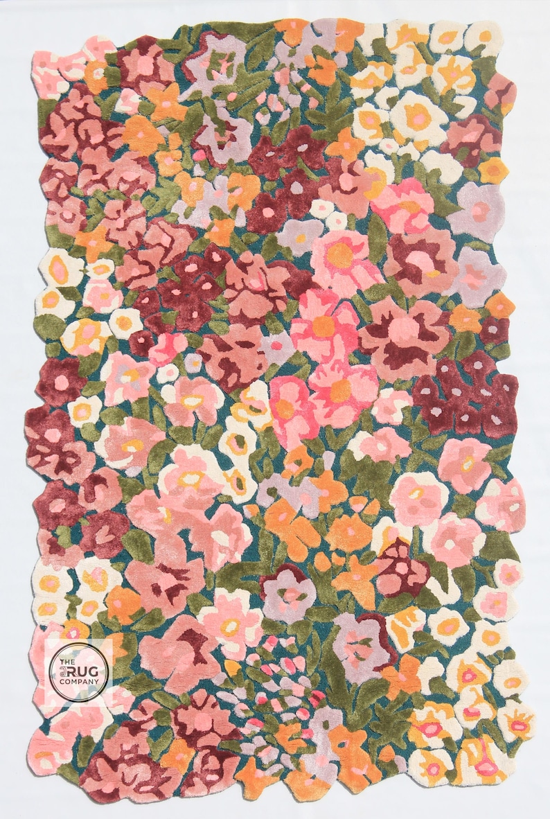 Pu&ograve; includere: Un tappeto floreale con un fitto motivo di fiori rosa, arancioni, bordeaux e bianchi su sfondo verde acqua. Il tappeto ha un bordo smerlato. Un logo &egrave; visibile nell'angolo in basso a sinistra.