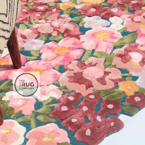 Pu&ograve; includere: Un tappeto floreale con sfondo turchese e un motivo di fiori rosa, rossi, gialli e bianchi. Il tappeto &egrave; accanto a una sedia con un motivo zebrato e gambe in legno. Il logo The Rug Company &egrave; visibile.