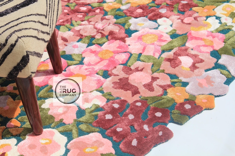 Pu&ograve; includere: Un tappeto floreale con sfondo turchese e un motivo di fiori rosa, rossi, gialli e bianchi. Il tappeto &egrave; accanto a una sedia con un motivo zebrato e gambe in legno. Il logo The Rug Company &egrave; visibile.