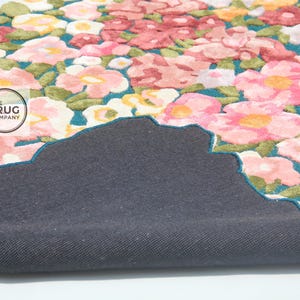 Pu&ograve; includere: Un tappeto floreale con un motivo di fiori rosa, rossi e gialli con foglie verdi su uno sfondo turchese. Il tappeto ha un retro grigio scuro e un bordo smerlato. Il logo The Rug Company &egrave; visibile.