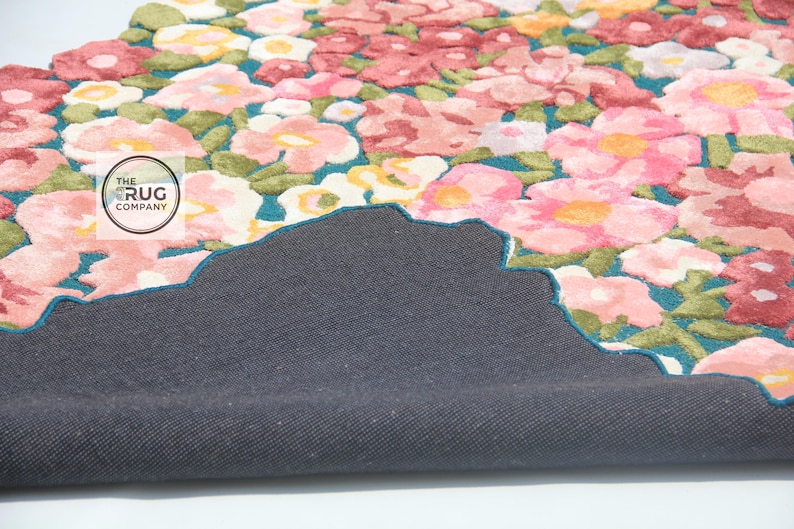 Pu&ograve; includere: Un tappeto floreale con un motivo di fiori rosa, rossi e gialli con foglie verdi su uno sfondo turchese. Il tappeto ha un retro grigio scuro e un bordo smerlato. Il logo The Rug Company &egrave; visibile.
