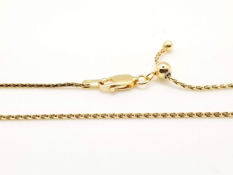 Puede incluir: Un collar dorado con un dise&ntilde;o de cadena de trigo. El collar presenta un cierre de mosquet&oacute;n y una cuenta ajustable en el extremo. La joya est&aacute; sobre un fondo blanco.