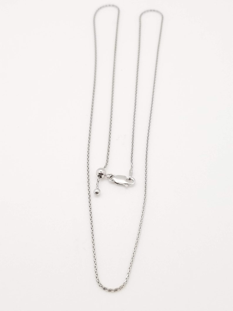 Puede incluir: Un collar ajustable plateado con una cadena delicada. El collar presenta una peque&ntilde;a bola y un cierre para ajustar la longitud. La cadena se muestra sobre un fondo blanco.