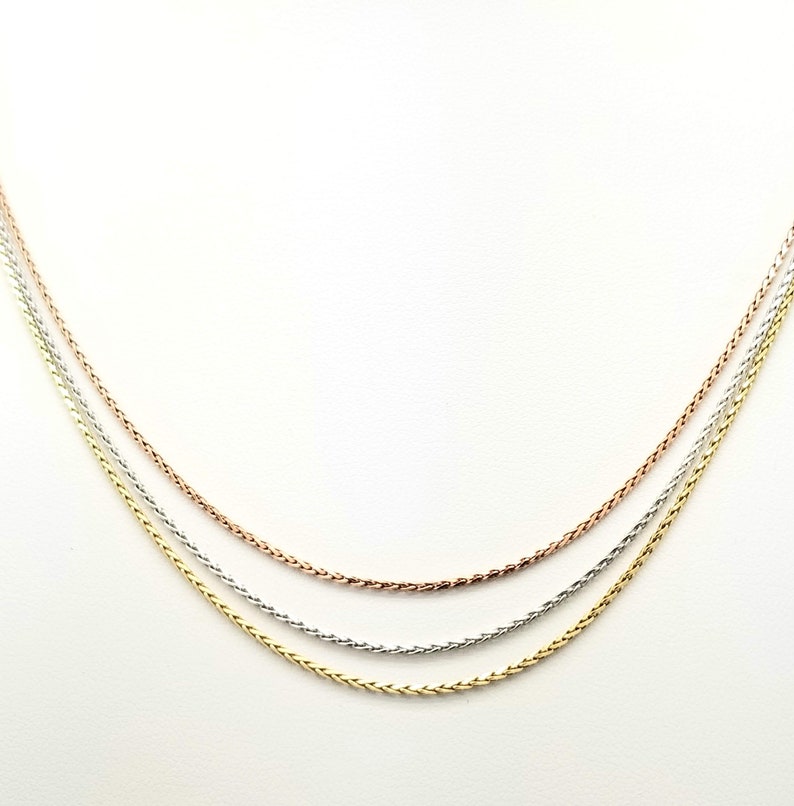 Puede incluir: Tres delicados collares en tonos oro, plata y oro rosa. Las cadenas tienen un dise&ntilde;o de trigo, cada una paralela a las otras sobre un fondo blanco. Los collares probablemente est&aacute;n hechos de metales preciosos.