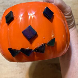 May include: An orange ceramic pumpkin with a jack-o'-lantern face made of dark, iridescent tiles. The pumpkin has a glossy finish and a rounded shape, perfect for Halloween decorations.
