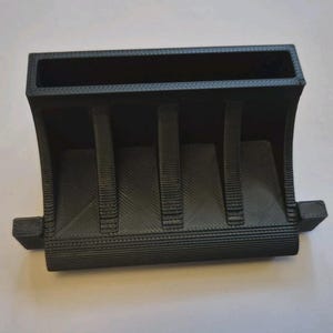 May include: A black, 3D-printed rectangular organizer featuring a wide top opening, three vertical support ribs along the front face, and two small rectangular tabs extending from the bottom sides. The surface displays a visible layered texture.