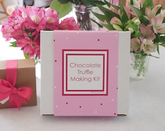 May include: A white box with a pink and white polka dot design. The box has a red border and the text "Chocolate Truffle Making Kit" on the front. The box is sitting on a white surface with pink flowers in the background.