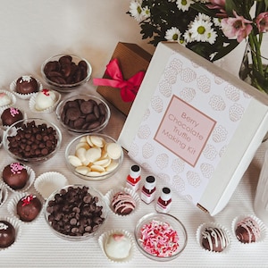 May include: A white box with a pink and white ribbon, labelled "Berry Chocolate Truffle Making Kit" with a pattern of strawberries. The box is surrounded by ingredients for making truffles, including chocolate chips, white chocolate chips, sprinkles, and chocolate truffles.