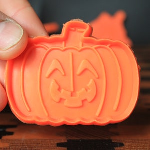May include: An orange plastic pumpkin-shaped cookie cutter with a carved jack-o'-lantern face. The cutter is held by a hand, with other cutters visible in the background. Perfect for Halloween-themed baking.
