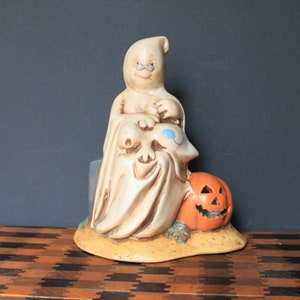 May include: A ceramic ghost figurine holding a small ghost in its arms, with a smiling jack-o'-lantern pumpkin at its feet. The ghost is white with blue eyes and a black mouth. The pumpkin is orange with a black face.