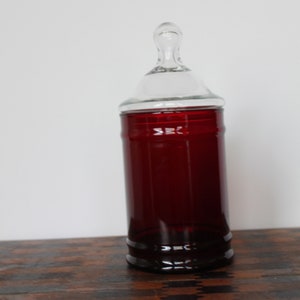 May include: A clear glass jar with a red glass base and a glass lid. The jar has a rounded top and a cylindrical body.