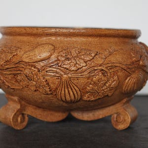 May include: A brown, handcrafted ceramic bowl with a detailed relief carving of gourds, leaves, and vines. The bowl has a rounded shape and sits on three curved feet, showcasing a rustic, earthy aesthetic.