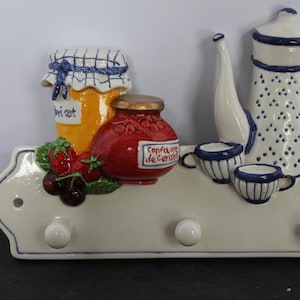 May include: A ceramic wall-mounted hook rack featuring a three-dimensional relief of a yellow jar labeled apricot, a red jar labeled confiture de cerises, a white teapot with blue polka dots, and two small cups. The base is cream with blue trim.