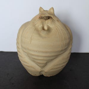 May include: A beige ceramic cat sculpture with a rounded body and a slightly flattened face. The cat has small ears and carved details, including vertical lines on its body. The sculpture is on a dark surface.